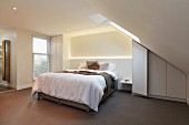 Upholstered bed in natural shades with indirect lighting in projecting masonry headboard and fitted wardrobe under sloping ceiling