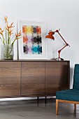 Retro Sideboard mit dunkler Holzfront, darauf Tischleuchte im Industriestil und Bild mit grafischem Motiv