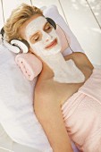 A blonde woman wearing headphones relaxing with a nourishing mask on her face, neck and chest