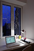 Laptop next to roll of paper and desk lamp on table below decorated window at twilight