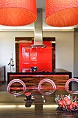 Orange lamps above dark exotic wood table and metal chairs with circular backrests in open-plan exotic wood kitchen with red fitted elements