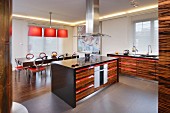 Cooker in island counter in open-plan kitchen with exotic wood fronts and red accents on handles; colour-coordinated dining area in background
