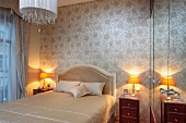 French bed with upholstered headboard and table lamps on bedside cabinets against wall with pale silk wallpaper in traditional bedroom