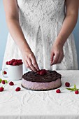 A woman placing fresh raspberries on a raspberry cheesecake with chocolate glaze
