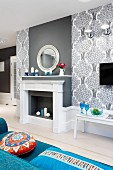 Elegant, grey and white faux fireplace and chimney breast flanked by wallpaper with pattern of stylised trees; colourful scatter cushions on blue sofa in foreground