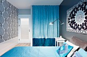 Wandteppich über Doppelbett mit hellblauen Textilien und zweifarbig blauer Vorhang vor einer Schranknische