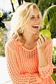 A blonde woman wearing an apricot, open-work jumper holding a green apple