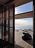 Blick durch offene Lamellentür auf Holzdeck mit Sitzsack und Sonnenliegen vor Meer