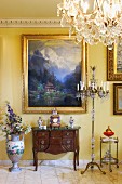 Antique, dark wood bureau below gilt-framed landscape on yellow-painted wall with chandelier floor lamp to one side