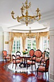 Gilt chandelier in front of period lounge area in semicircular window bay; chairs with red and gold ornamental pattern on upholstery in grand interior