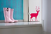 Pink child's wellingtons on platform next to white-painted wooden door with red stag motif