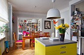 Free-standing, bright yellow counter with drawers and grey worksurface, vase of narcissus, Easter decoration hanging from pendant lamp and Biedermeier-style dining set in open-plan interior
