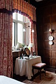Dressing table below window with toile-de-jouy curtains and decorative china wall plates on wood-panelled walls in bedroom