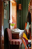 Stuhl mit gestreiftem Bezug vor dunkelgrün gestreifter Badezimmer-Tapete, Waschtisch im Antikstil vor Fenster