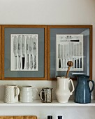 Collection of jugs on shelf below framed pictures of knives on wall