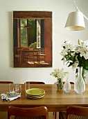 Stacked plates, glass vases and bouquet of white lilies on oak dining table in front of painting on wall