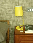 Yellow, retro bedside lamp on wooden bedside cabinet against wallpapered wall