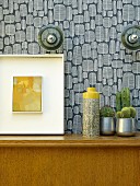 Vase next to cacti and picture on wooden cabinet against wall with retro wallpaper
