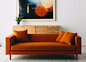 Orange sofa with cushions of the same color in front of a sideboard made of precious wood and a framed picture in the background