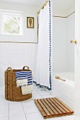 Rustic laundry basket with blue and white striped towels on tiled floor in front of bathtub with white shower curtain