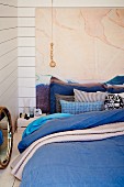 Matratzenlager auf Boden mit blauer Bettwäsche, Kissenstapel vor Gemälde an weisser Holzwand als Bettkopfteil