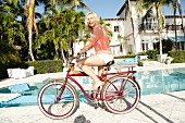 A blonde woman on a bike by a hotel pool wearing and apricot openwork jump and white shorts