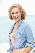 A blonde woman by the sea wearing an apricot top and a denim shirt