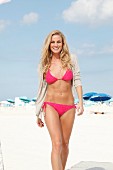 Blonde Frau mit pinkfarbenem Bikini und hellem Jäckchen geht am Strand entlang