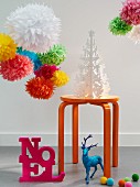 Colourful paper pom-poms hung over a white paper Christmas tree on a stall with a 'Noel' decoration and a neon blue deer on the floor
