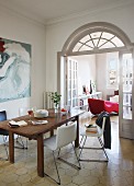 Wooden table and modern chairs with shell seats in front of open double doors with arched transom window and view of red couch in living room