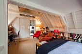 Multifunctional attic room with home office, seating area, bookcases and bed under exposed, white-painted wooden roof structure