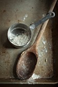 A wooden spoon and a measuring cup with flour on a baking tray