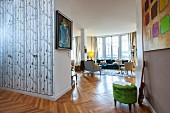 Tree-trunk-patterned wallpaper, green velvet pouffe in open doorway and lounge with glass wall in interior with continuous herringbone parquet floor