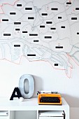 Stilisierter Stadtplan mit Nägeln und Fäden bespannt dargestellt als Wanddekoration, orangefarbene Retro Schreibmaschine und Deko-Buchstaben auf weißem Regal