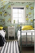 Vintage-style bedroom with floral wallpaper and bedside cabinet between twin metal beds with frames painted grey below window