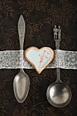 A heart-shaped biscuit decorated with egg white glaze and a stamped motif between wedding-themed spoons