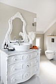 Modern bathroom with sloping ceiling and wall-mounted toilet; vintage chest of drawers painted white with round, modern countertop washbasin and carved mirror