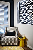 Reading armchair with graphic upholstery in corner next to retro side table and wall aperture filled with geometric window grate