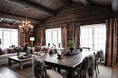 Offener Wohnraum in rustikalem Holzhaus, graues Fell auf Stühlen um Massivholztisch, im Hintergrund gemütliche Loungebereich