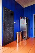 Schiefertafel an Tür, Abfalleimer und Edelstahl Kühlschrankkombination in einer Küche mit blau lackierter Holzwand