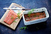 Graved lax being made: marinated salmon fillets in a rectangular container with spices and dill and on a chopping board