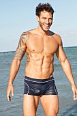 A young, tattooed man by the sea wearing bathing trunks