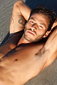 A young, topless, tattooed man lying on a sandy beach