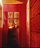 Metallvase mit Bambuszweigen und Windlichter auf zylindrisch geformter Kommode, vor rot getönter Wand