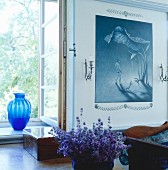 Bowl of purple flowers on table in front of window with blue glass vase on windowsill; painting in shades of blue to one side