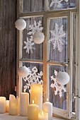 Lit, white pillar candles on sill of window decorated with festive paper snowflakes
