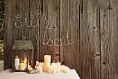 Brennende Kerzen auf Tisch und in Laterne vor Bretterwand mit weihnachtlicher Botschaft