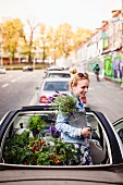 Urban gardening: young woman in convertible car with foliage plants and vegetable plants