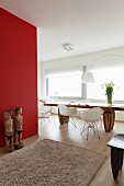 Pale grey Berber rug on wooden floor, wooden sculptures against red wall and seating area with white, classic chairs in seating area