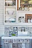 Rustic sink unit with wooden base cabinet and splashback made from Indonesian tiles below shelving on white, wood-clad wall